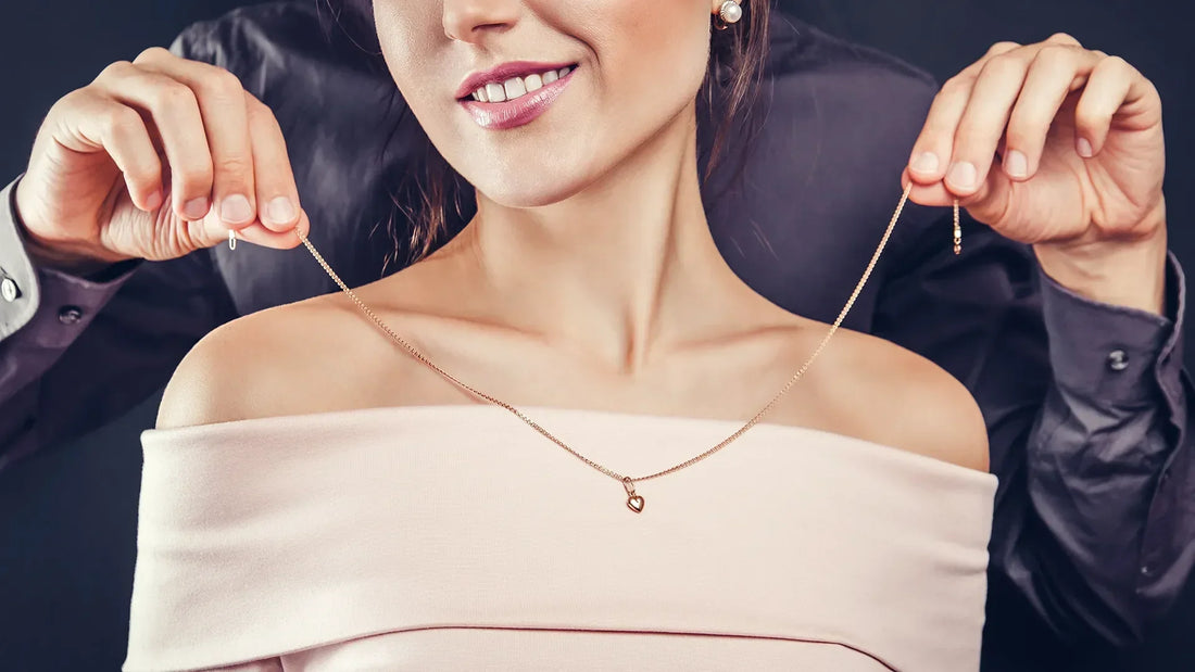 Junge Frau in rosa schulterfreiem Oberteil hält lächelnd die Enden einer feinen Goldkette mit kleinem Herzanhänger hoch