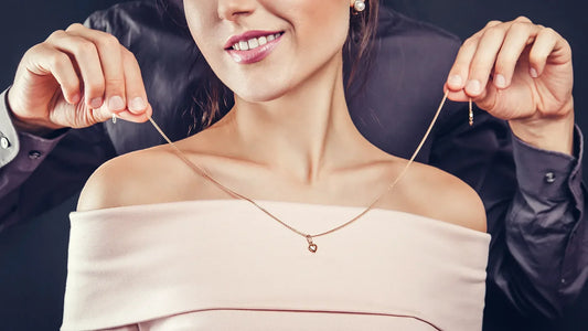 Junge Frau in rosa schulterfreiem Oberteil hält lächelnd die Enden einer feinen Goldkette mit kleinem Herzanhänger hoch
