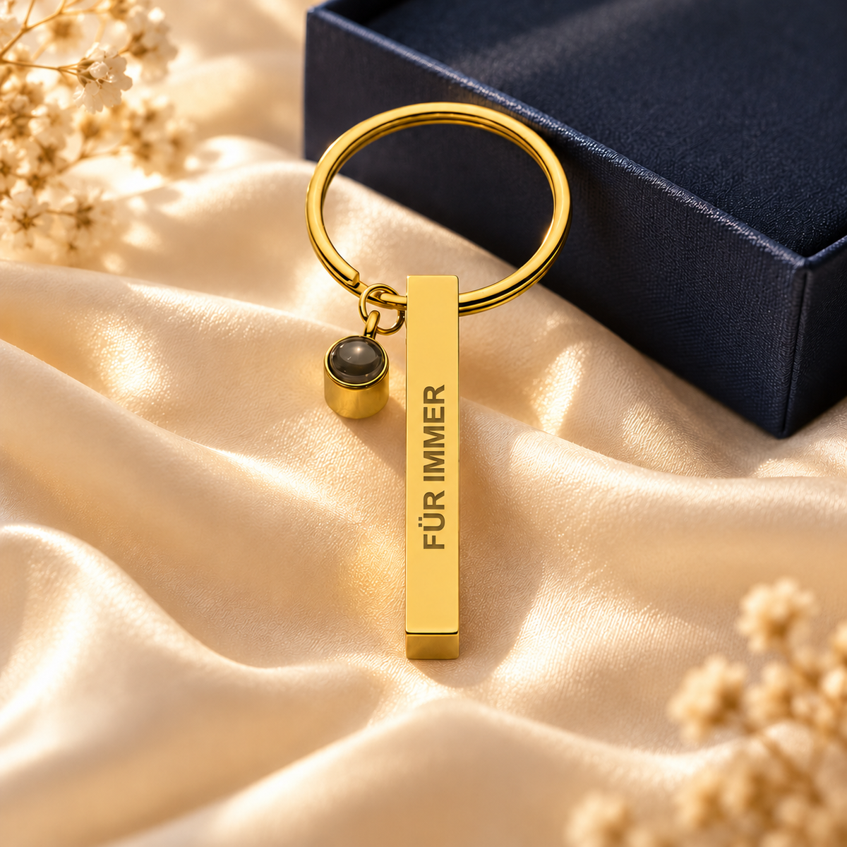 Schlüsselanhänger mit Gravur „Für immer“ in Gold mit Fotolinse vor einer Geschenkbox auf heller Seide
