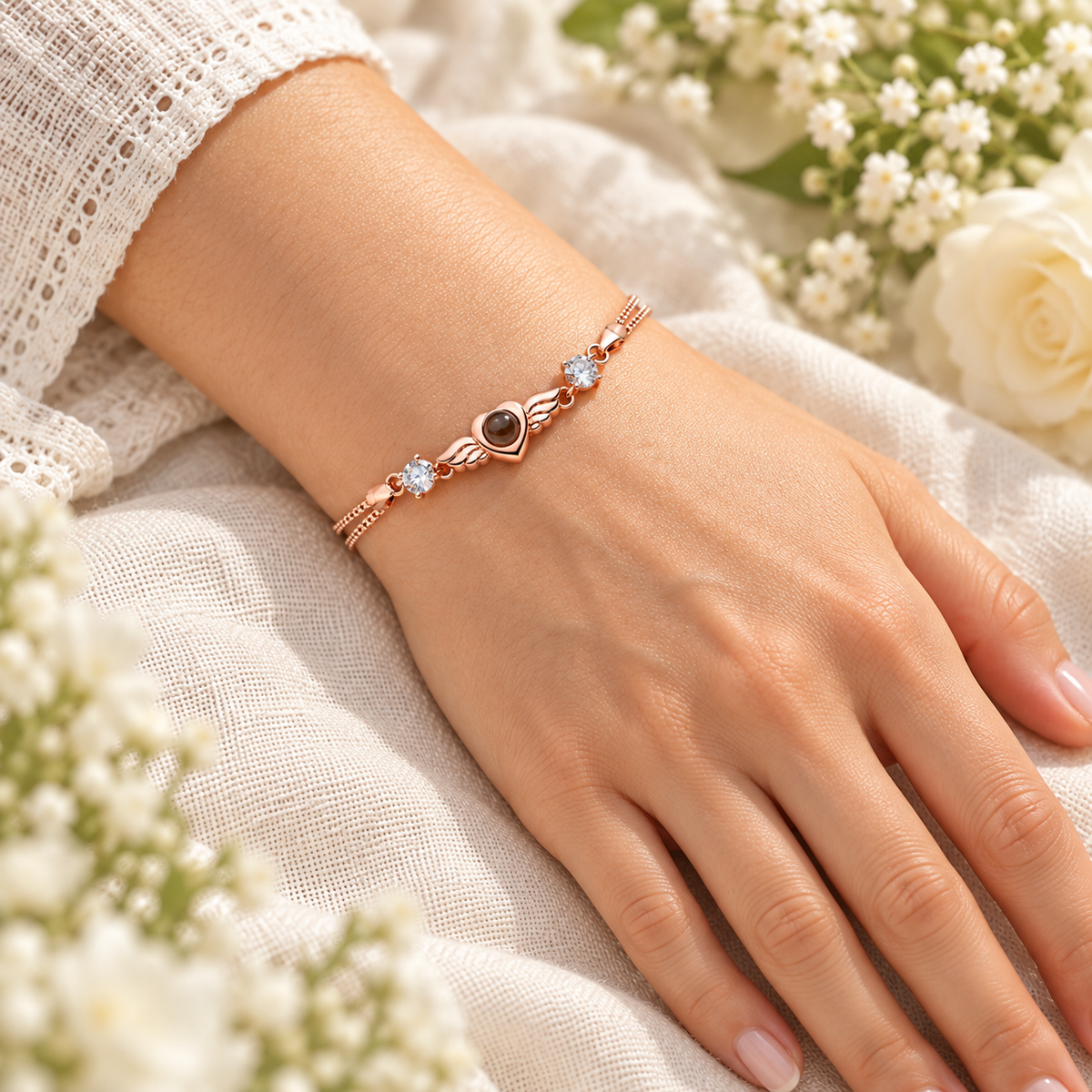 Roségoldfarbenes Flügelherz-Fotoarmband von 4MyLove mit Fotolinse und zwei klaren Schmucksteinen, getragen an einer Hand auf hellem Leinen