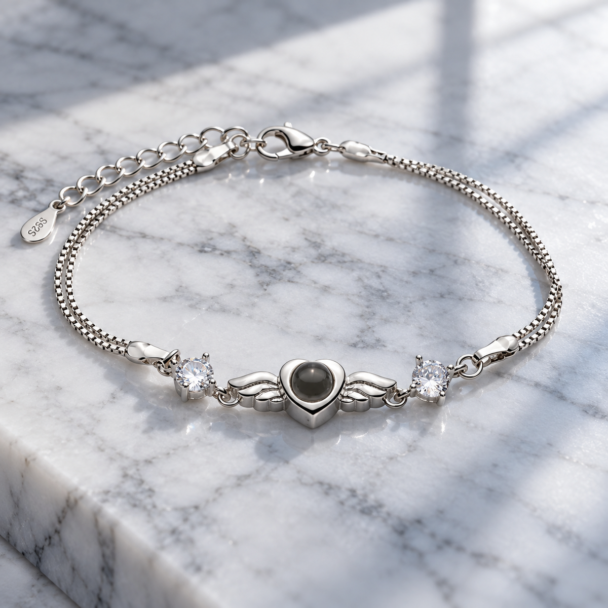Silberfarbenes Fotoarmband von 4MyLove mit herzförmiger Fotolinse, Flügelmotiv und zwei klaren Schmucksteinen auf hellem Marmor